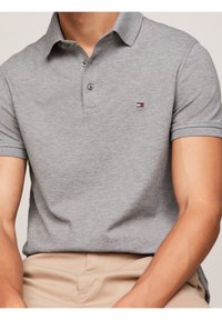 Homme portant un polo à manches courtes gris clair avec un petit logo rectangulaire rouge, blanc et bleu sur la poitrine, et un pantalon beige.