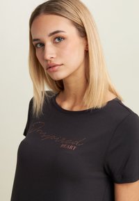 T-shirt nera a maniche corte, con la scritta in corsivo "Inspired" e "HEART" in decorazioni oro rosato sul davanti. Tessuto di cotone morbido.
