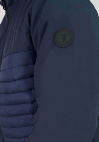 Marineblauwe jacket met een gequiltte textuur op het lijf, gladde stof op de mouwen en een ronde zwarte logopatch op de linkermouw.