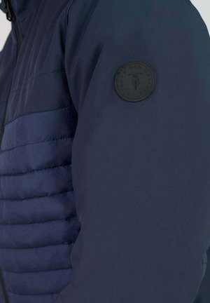 Veste marine avec une texture matelassée sur le torse, un tissu lisse sur les bras et un patch logo noir rond sur la manche gauche.