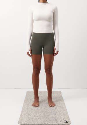 Femme debout pieds nus sur un tapis de yoga Nike gris clair, portant un haut blanc moulant à manches longues et un short Nike vert olive, sur fond blanc.