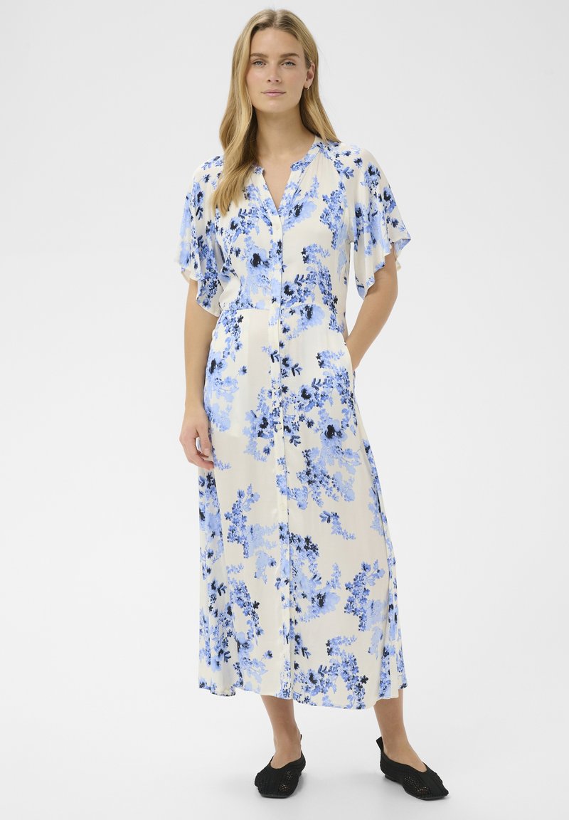 Donna che indossa un vestito midi color crema con stampa floreale blu, maniche svolazzanti, chiusura con bottoni sul davanti e scarpe nere basse, in piedi con una mano in tasca.