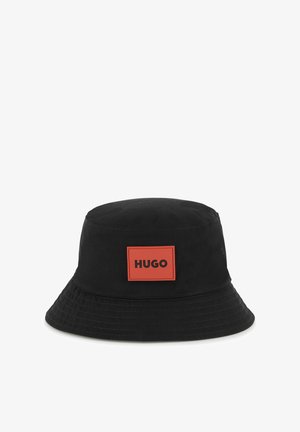 Cappello a secchiello nero in tessuto, con una visiera larga e piatta. Un'etichetta rettangolare arancione con "HUGO" in nero è ben visibile sulla parte anteriore.