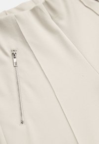 Zbliżenie beżowej tkaniny z pionowym srebrnym zamkiem błyskawicznym oraz nachodzącymi na siebie przeszytymi panelami tworzącymi strukturalny wzór.