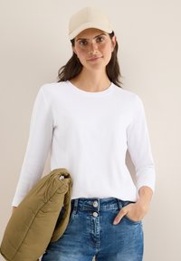 Valkoinen pitkähiha-silkkipaita pyöreällä kaula-aukolla, yhdistettynä sinisiin farkkucaprihousuihin. Mukana on myös beige lippalakki ja oliivinvihreä laukku.