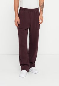 Burgunderfarvede sweatpants med en afslappet pasform, der har to rummelige forlommer og en elastisk talje, båret med hvide sneakers.