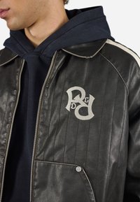 Zwarte lederen jas met verticale stiksels, zilveren accenten en een crèmekleurig geborduurd logo op de linker borst. Draagt over een marineblauwe hoodie.