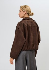 Ruda dirbtinės zomšos striukė su per dideliu dizainu, nuleidžiamos pečių linijos ir elastinga apacia. Ypatybės: didelis apykaklė ir užtrauktukas.