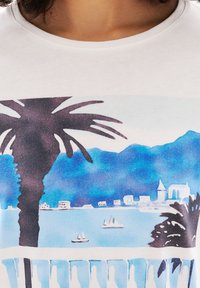 Valkoinen t-paita, jossa on värikäs akvarelliprintti, joka esittää palmupuun, vuoria ja veneitä sinisellä merellä.