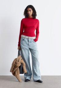 Rotes Langarmshirt, kombiniert mit hellblauen weit geschnittenen Jeans, einem cremefarbenen Gürtel und einer schwarzen Handtasche, über die ein gestrickter beiger Pullover drapiert ist.