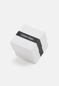 Biały kartonowy pudełko z czarną opaską z napisem "Calvin Klein" w białym kolorze. Pudełko ma geometryczny wzór i gładką powierzchnię.