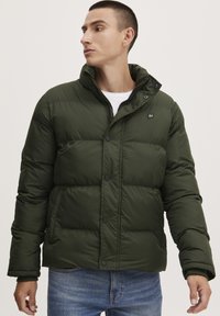 Green puffer jacket with a high collar, zip and snap button closure, and quilted design. Worn over a white shirt, paired with blue jeans.
