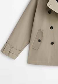 Beigenvärinen trenssitakin hiha ja etuosa mustilla napeilla, viistolla läppätaskulla ja säädettävällä hihansuun remmillä valkoisella taustalla.