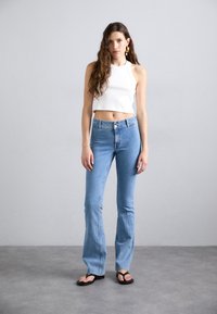 Top corto bianco con scollatura rotonda, abbinato a jeans a zampa di elefante azzurri chiari. Modella con ciabatte nere, in piedi su uno sfondo grigio.