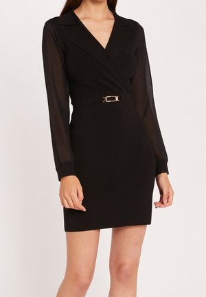 Kobieta w czarnej mini sukience z długim rękawem, przezroczystymi rękawami i złotym paskiem z klamrą w talii.