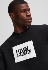 Hanorac negru cu un logo alb dreptunghiular proeminent, având inscripția "KARL LAGERFELD" cu font bold negru. Textură netedă și decolteu rotund.