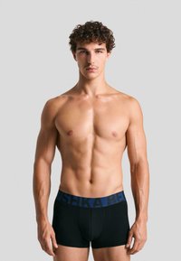 Boxeri negri cu un brâu albastru care prezintă numele brandului. Materialul pare neted și elastic, aderând strâns la talie și coapse.