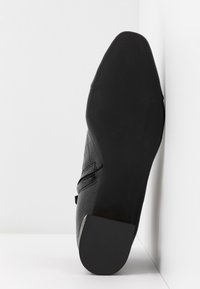Bottine en cuir noir avec semelle texturée et fermeture éclair latérale, positionnée debout contre un coin blanc entre le mur et le sol.