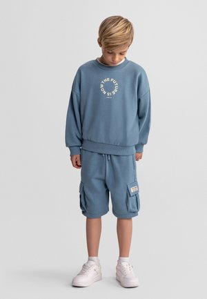 Blond chłopiec w niebieskiej bluzie z napisem „THE FUTURE IS NOW”, dopasowanych spodenkach bojówkach, białych skarpetach i białych trampkach, patrzący w dół.