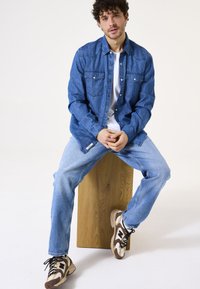 Donkerblauwe denim shirt, met knopen en twee borstzakken, gecombineerd met lichtblauwe jeans en sneakers met bruine accenten op een houten kruk.