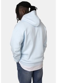 Felpa azzurra chiara con un grande cappuccio, realizzata in materiale morbido, con polsini e orlo a costine. Indossata sopra una camicia bianca, con jeans dalla vestibilità comoda.