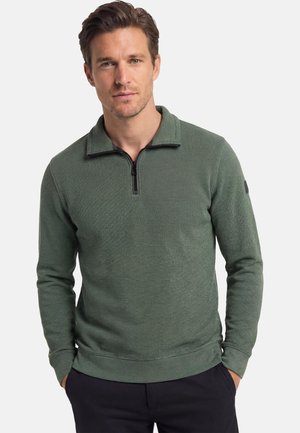 Mand iført en grøn langærmet pullover med kvart lynlås, med hænderne i lommerne, vendende frem mod en ensfarvet baggrund.