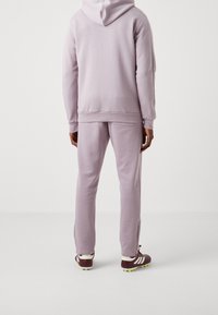 Sudadera con capucha lila y pantalones deportivos a juego hechos de tela suave. La sudadera tiene cordón, y los pantalones cuentan con bolsillos laterales y cremalleras en los tobillos.