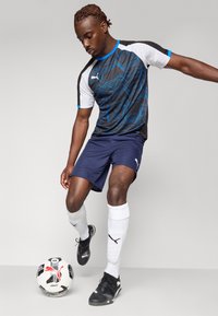 Zwart-wit atletisch shirt met een blauw golvend patroon, gecombineerd met marineblauwe shorts. Speler draagt zwarte voetbalschoenen en witte sokken, staand met een voetbal.