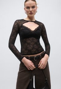 Μαύρο μακρυμάνικο τοπ από δαντέλα με λαιμόκοψη με κυματιστό σχέδιο και άνοιγμα κατσαβιδόσχημο. Η υφή είναι περίπλοκη με λουλουδένια μοτίβα.