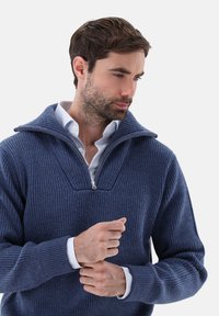 Marineblauer gerippter Pullover mit einem halben Reißverschluss und V-Ausschnitt. Über einem weißen Hemd mit Kragen gestylt, das die Strickstruktur zur Geltung bringt.