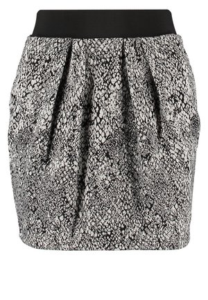 Femme portant un haut à manches longues blanc, une jupe à motif noir et blanc, des collants noirs, des bottines noires et un sac bandoulière noir, debout avec une main sur la hanche.