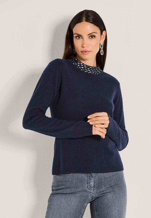 Frau mit langen dunklen Haaren, die einen dunkelblauen Pullover mit einem gemusterten hohen Kragen und hellgraue Jeans trägt, steht vor einer schlichten Wand.