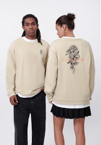 Beige oversized sweatshirt with a front logo and large rose and snake graphic on the back. Features ribbed cuffs and a textured fabric.
