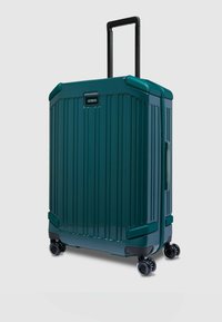 Een blauwgroene harde koffer met verticale ribbels, uitschuifbare zwarte handgreep en vier zwenkwieltjes voor mobiliteit. Voorzien van een zijkant handgreep.