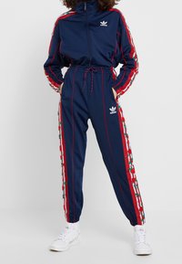 Person som bär en marinblå Adidas träningsoverall med röda och vita blommönstrade ränder och vita sneakers, stående med händerna i fickorna.