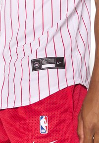 Biała koszulka baseballowa z czerwonymi pionowymi paskami, z logo marki i guzikami, w zestawie z czerwonymi siateczkowymi szortami z logo NBA.