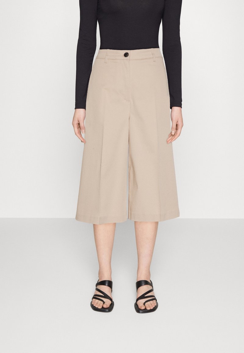 Personne portant un pantalon beige large et court, un haut noir à manches longues et des sandales noires à brides, debout devant un fond uni.