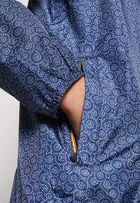 Blå mønstret jakke med elastiske manchetter, der har en lomme med en flerfarvet lynlås og en let, glat tekstur.