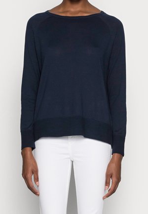 Persoon draagt een effen donkerblauwe trui met lange mouwen gecombineerd met een witte broek, staand tegen een neutrale achtergrond.