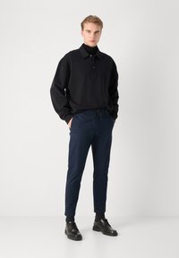 Black long-sleeve polo with a collar, paired with navy tapered trousers. Model wears black shoes. Smooth fabric, minimalist design.