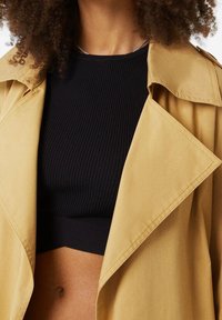 Un trench-coat beige clair avec un large col et des revers, superposé sur un crop top noir à côtes, mettant en valeur une silhouette ajustée et un contraste de textures.