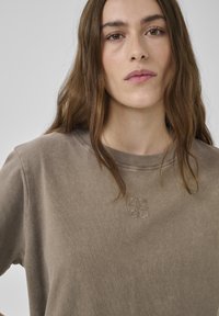 Junge Frau mit langen braunen Haaren trägt ein taupefarbenes Sweatshirt mit rundem Ausschnitt und einem dezenten bestickten Muster auf der Brust, neutraler Hintergrund.