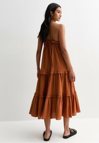 Robe maxi à volants de couleur marron, avec des bretelles fines et réglables, et un dos noué. Portée avec des sandales plates noires.