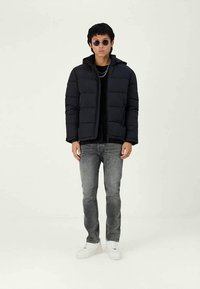 Jovem vestindo uma jaqueta puffer preta, jeans cinza, tênis brancos, óculos de sol e correntes de prata em camadas, posando em fundo branco.