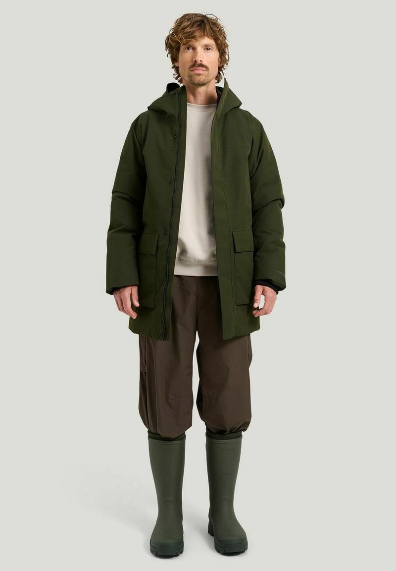 Man som bär en grön huva parka, beige sweatshirt, bruna byxor och gröna gummistövlar, står mot en ljus enhetlig bakgrund.