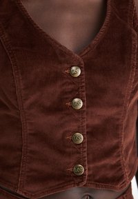 Bruine corduroy cropped top met een V-hals, voorzien van vijf messingen knopen met "Lee" erop gedrukt, en een gestructureerd oppervlak.