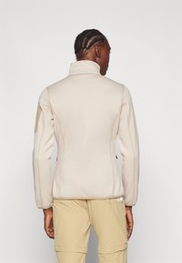 Veste en polaire beige avec un col montant, surface texturée, coutures contrastantes et renforts au niveau des coudes. Présente un design ajusté avec deux poches latérales.