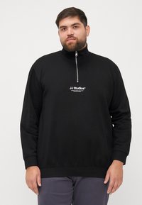 Sudadera negra con cremallera en el cuello, que presenta un estampado de logo en la parte delantera, mangas largas, puños acanalados y un ajuste relajado. Elaborada con una tela suave y texturizada.