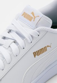 Bílé kožené tenisky s plochými bílými tkaničkami, zlatým logem Puma na boku a jazyku, a texturovanými detaily na svršku a podrážce.