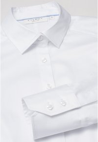 Chemise blanche boutonnée avec col classique, présentant une texture lisse, des boutons en perle et une coupe ajustée. Étiquette visible à l'intérieur du col.